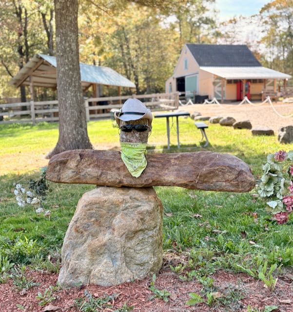 IMG_2363 Rock sculpture wearing sunglasses, hat, and bandana in a rural setting with barn and trees.