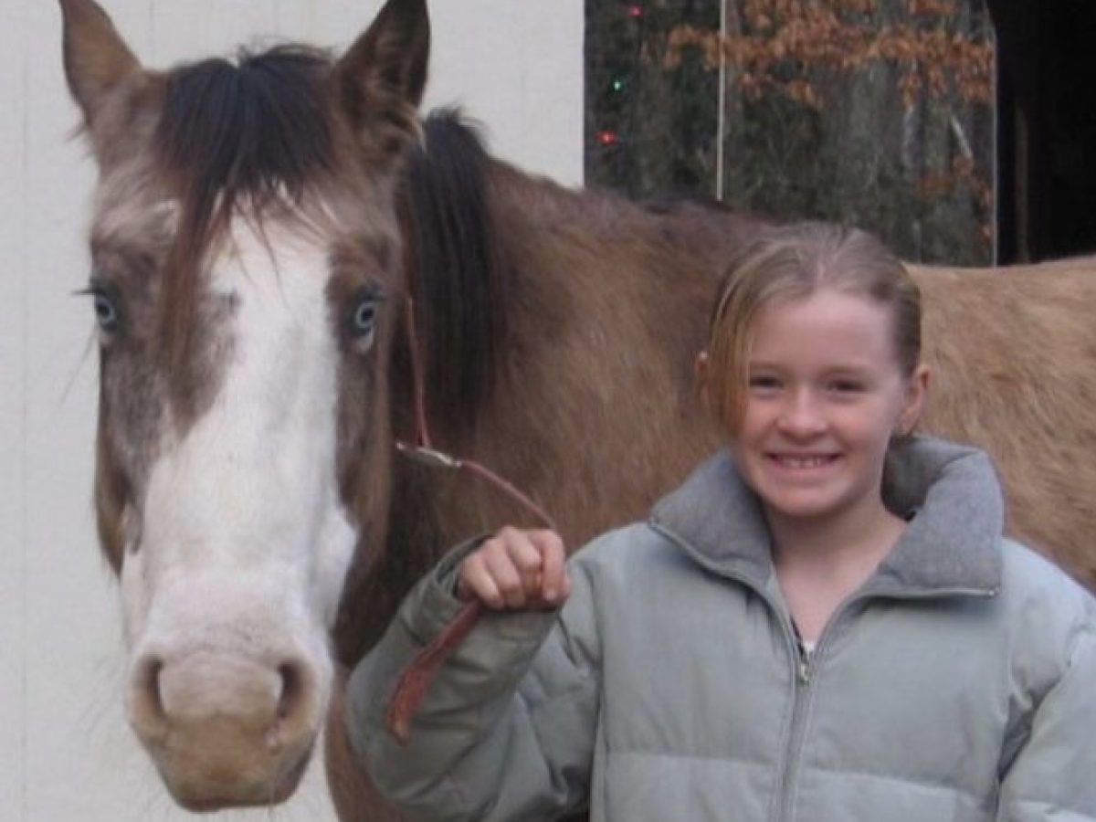 Smiling child in a gray jacket holding a rope with a brown horse outside a building.