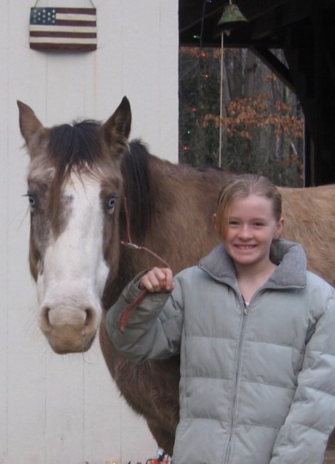 Smiling child in a gray jacket holding a rope with a brown horse outside a building.