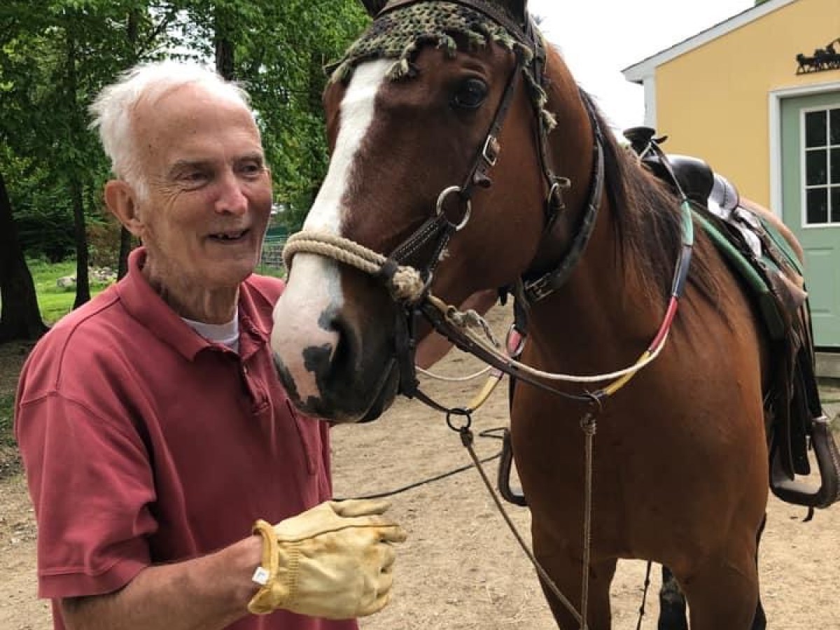 Elderly man smiling beside a brown horse in a saddle on a dirt path.