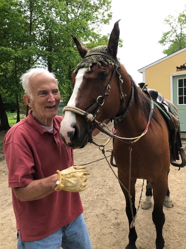 Elderly man smiling beside a brown horse in a saddle on a dirt path.