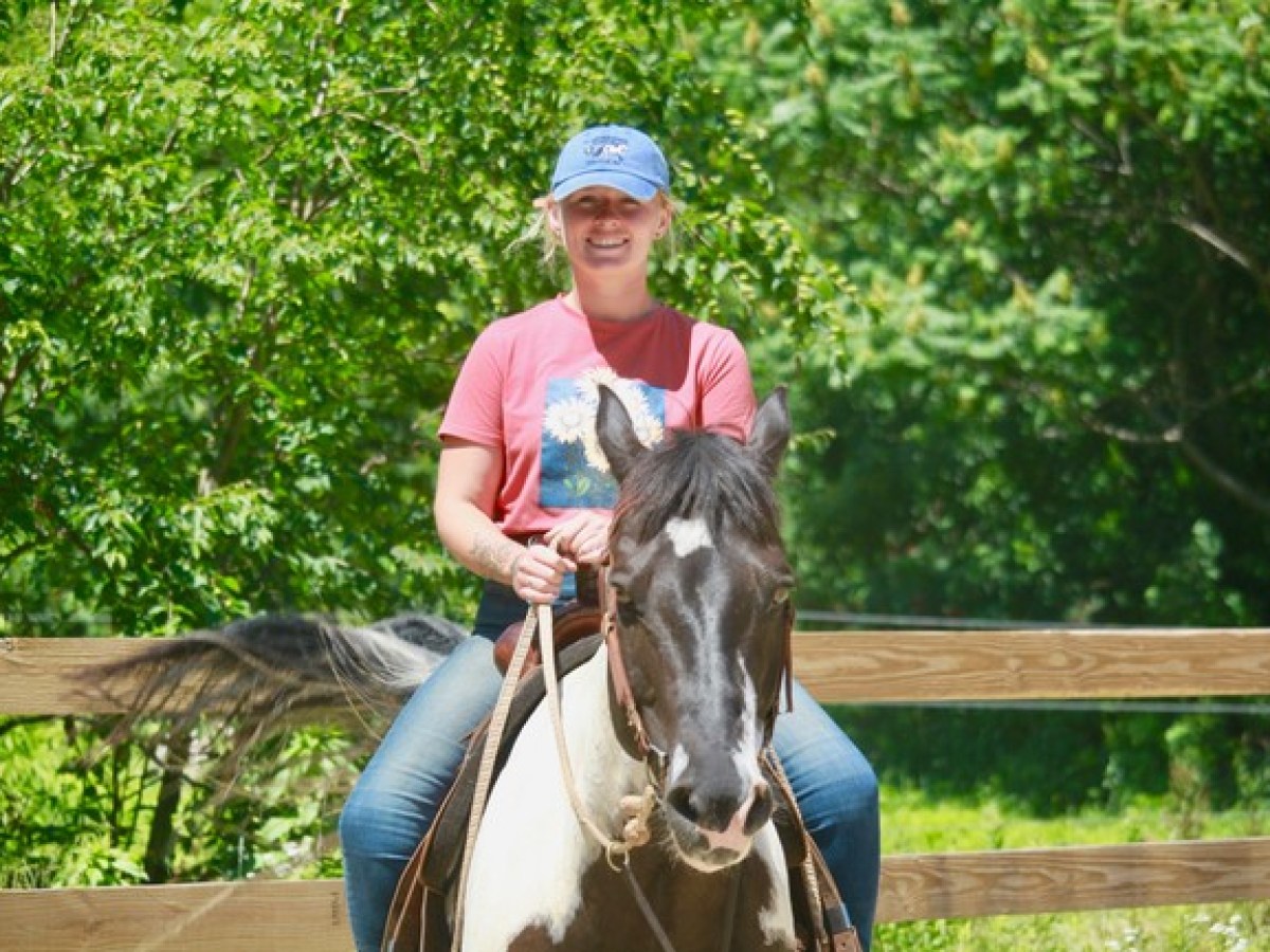 Person in red shirt and blue cap riding a black and white horse in a fenced area with trees.