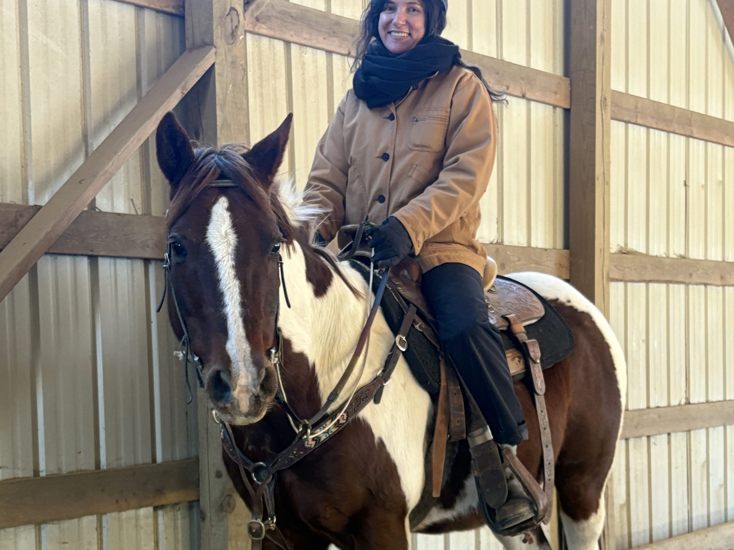 Person in a brown coat and helmet riding a brown and white horse indoors.