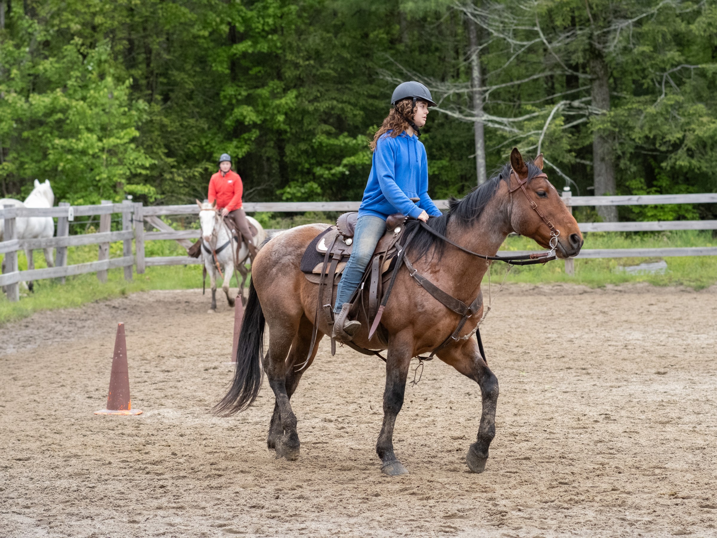 Two people riding horses in an outdoor fenced arena with trees in the background.