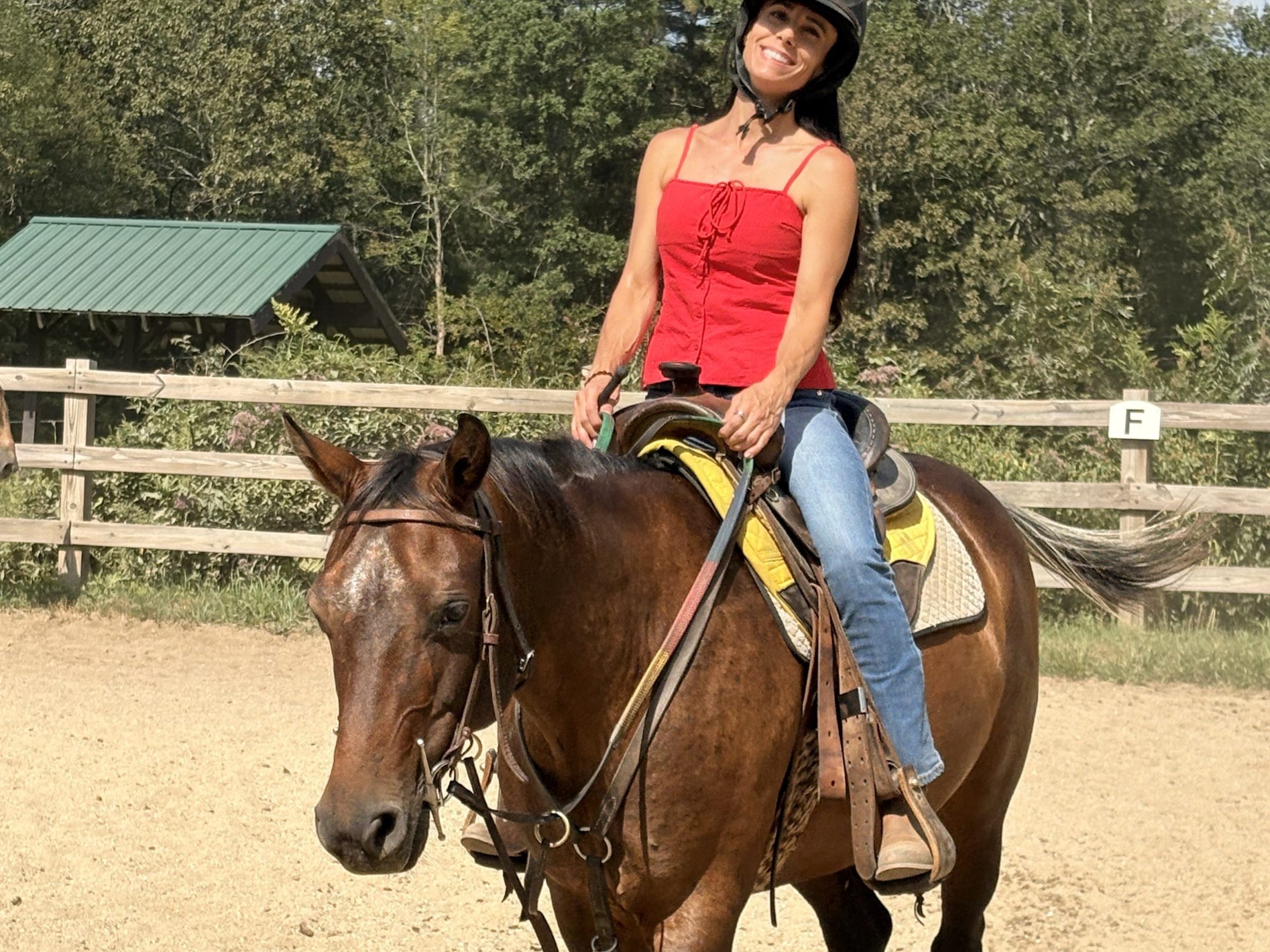Woman in red top riding a brown horse in an outdoor arena.