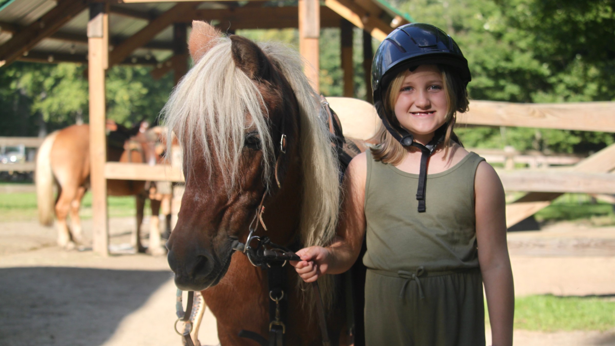 Child in helmet stands beside a pony in a fenced area under a wooden structure.