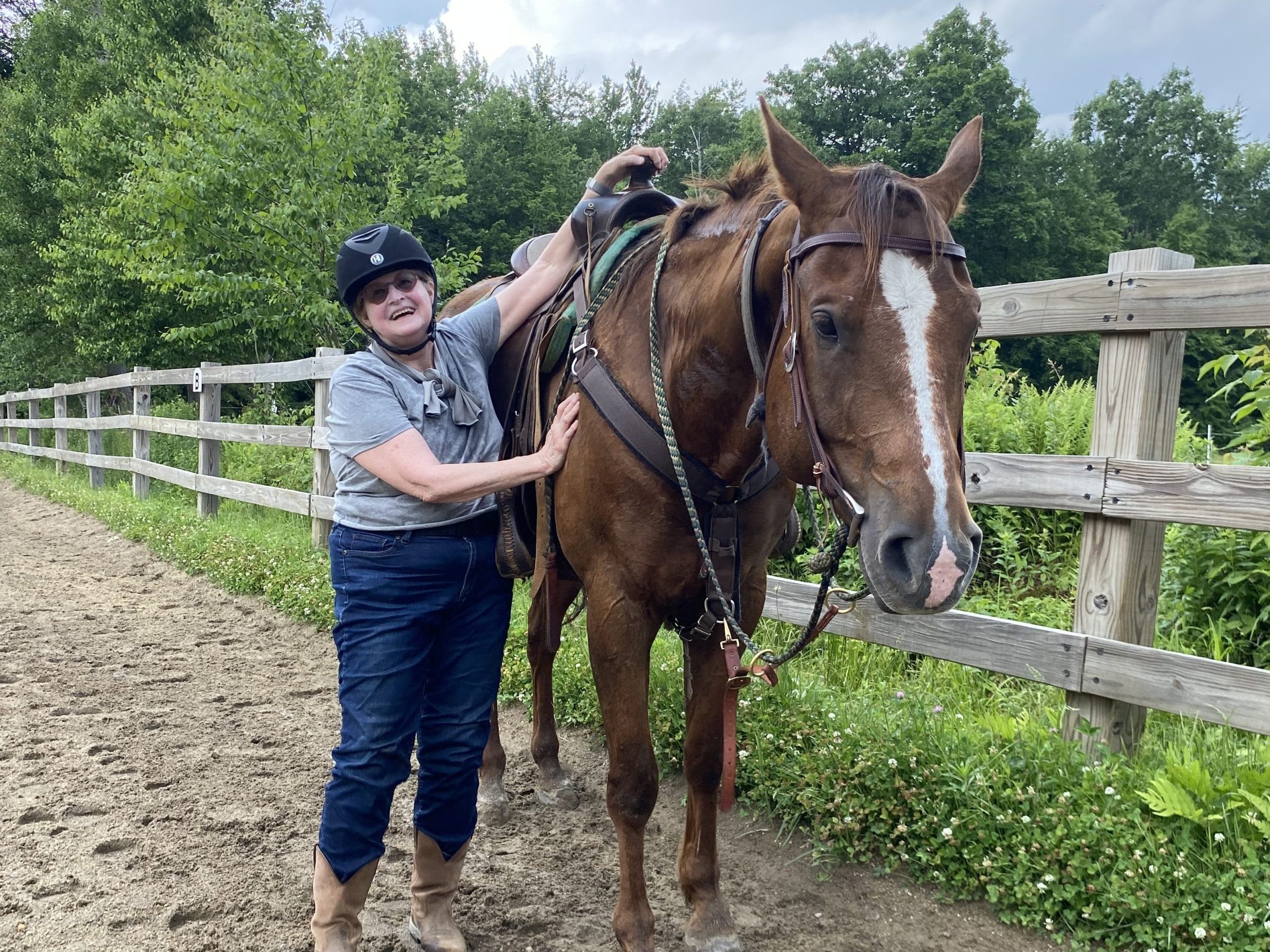 Person in riding gear standing beside a brown horse on a dirt path, with trees and a wooden fence in the background.