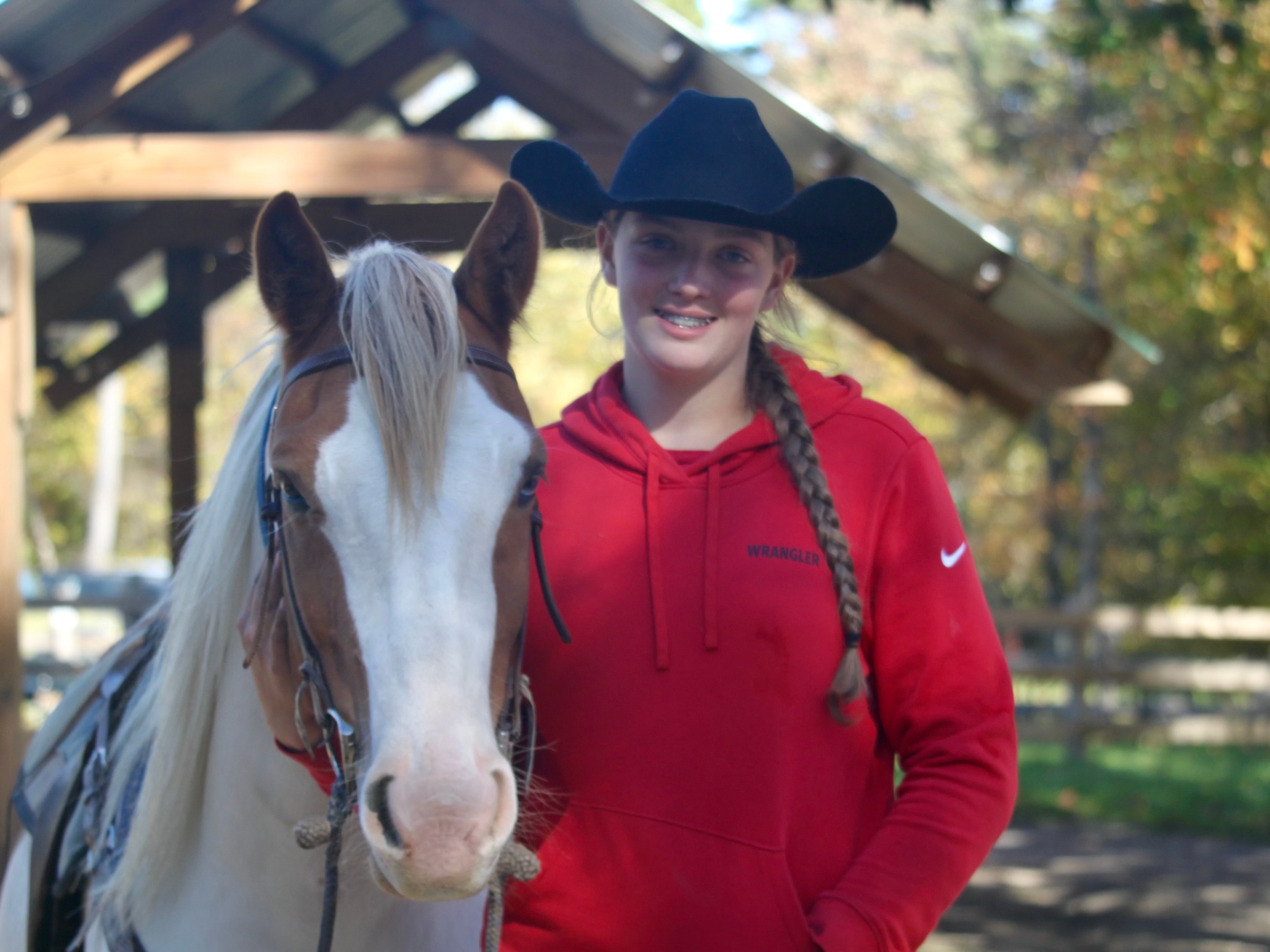 Person in red hoodie and black cowboy hat standing next to a horse with a bridle outside.