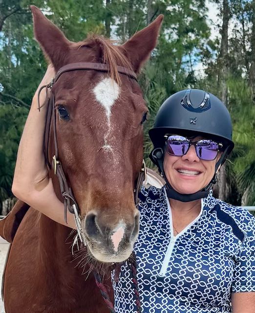Person with sunglasses and helmet smiling next to a brown horse.