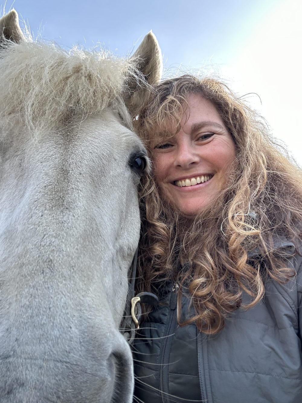 Person with curly hair smiling next to a white horse.